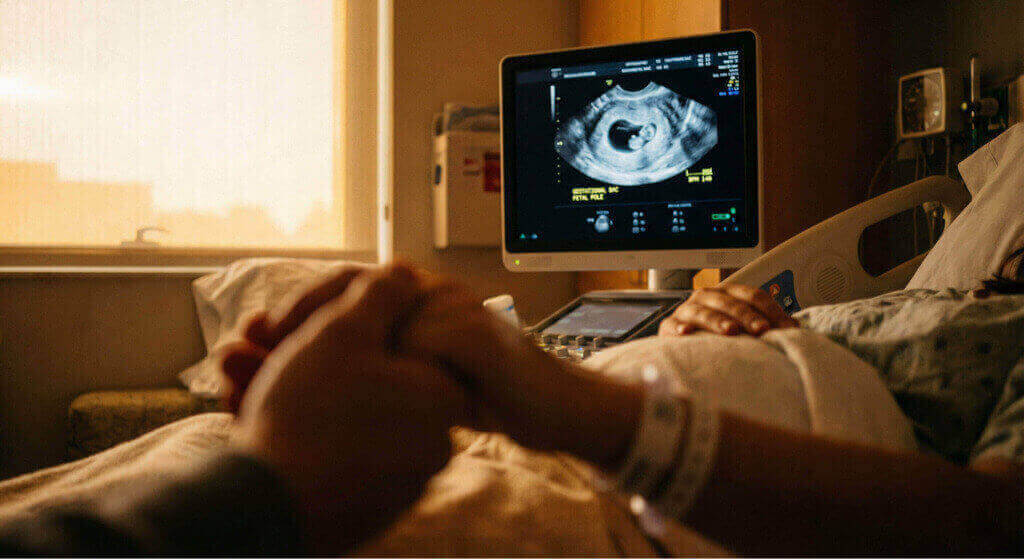 Ultraschallmonitor mit Fruchtsack und Embryopole, im Vordergrund eine stützende Hand